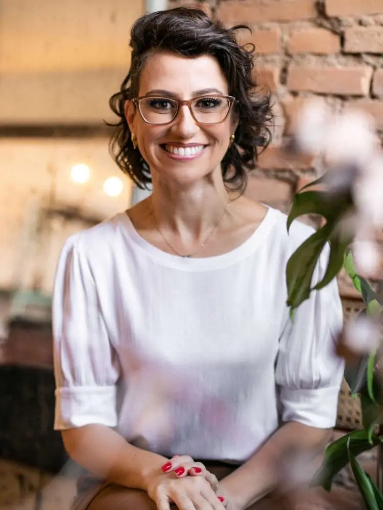 Mulher não-modelo com mais de 40 anos em ensaio fotográfico, usando blusa branca, óculos, sorrindo