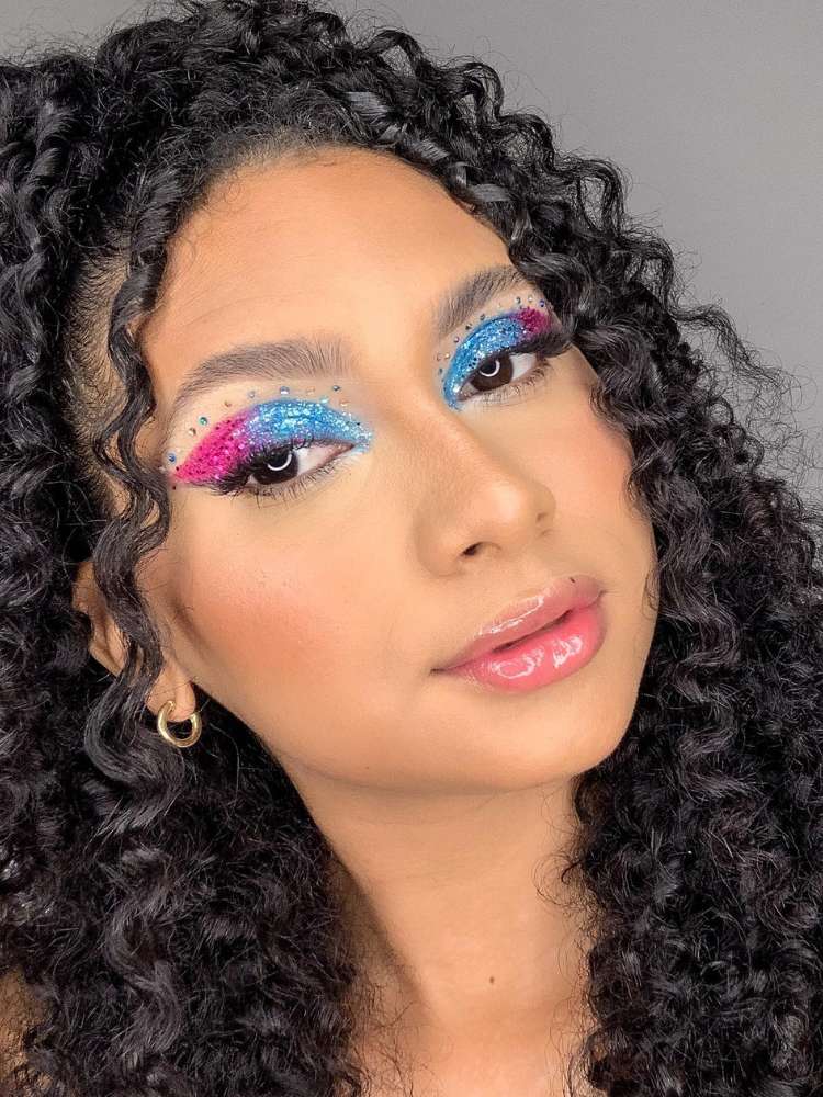 Mulher de pele morena com cabelo escuro cacheado iusando sombra azul e roxa nos olhos, com glitter e strass. 