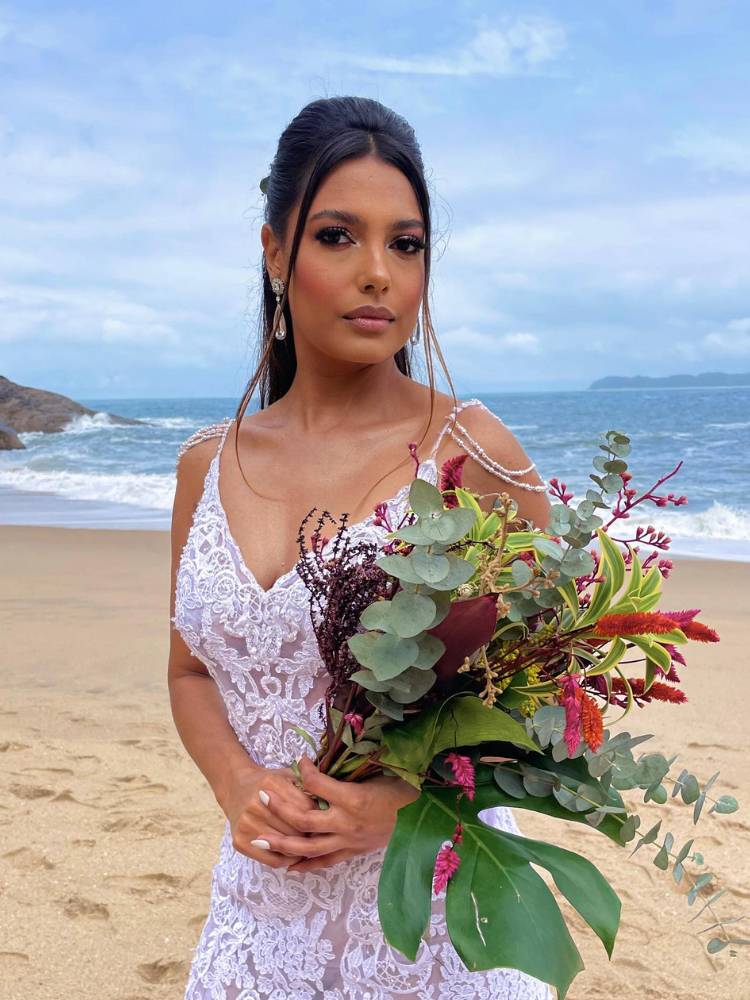 Noiva de pele negra na praia, usando vestido rendado de alça, penteado semi preso, maquiagem natural e segurando buquê colorido