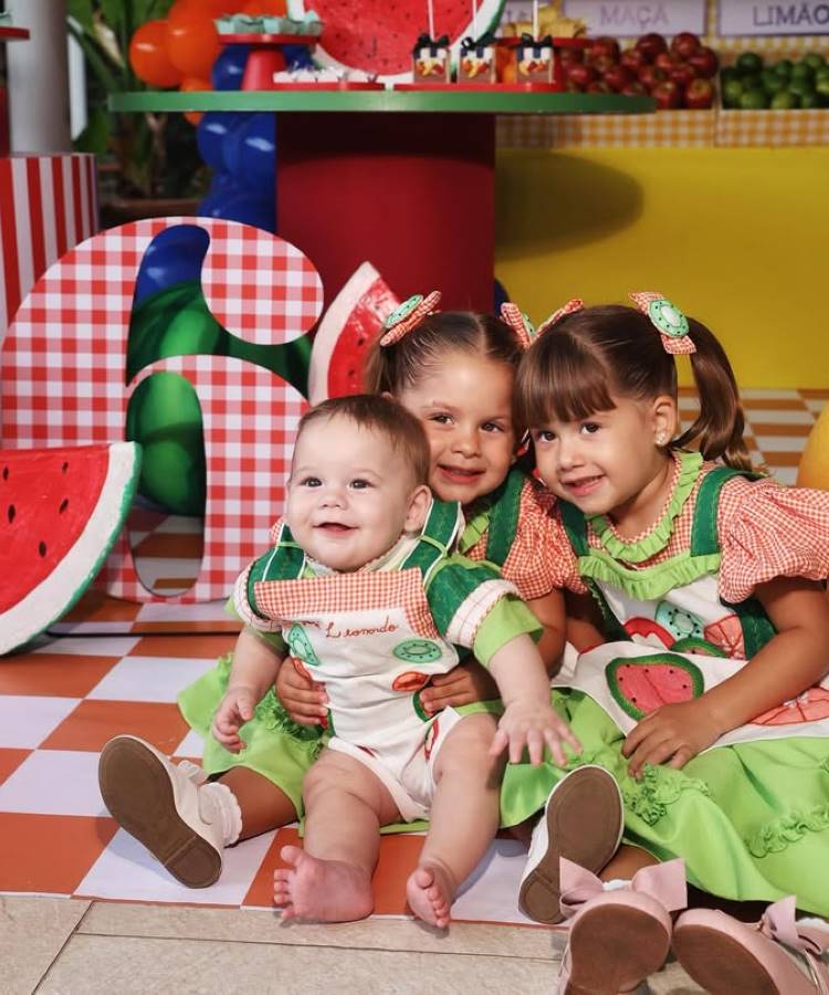 Três crianças sorrindo em uma festa temática de melancia, rodeadas por elementos decorativos coloridos. As duas meninas usam vestidos verdes com detalhes de xadrez laranja, enquanto o bebê veste um macacão temático, sentado à frente das irmãs.






