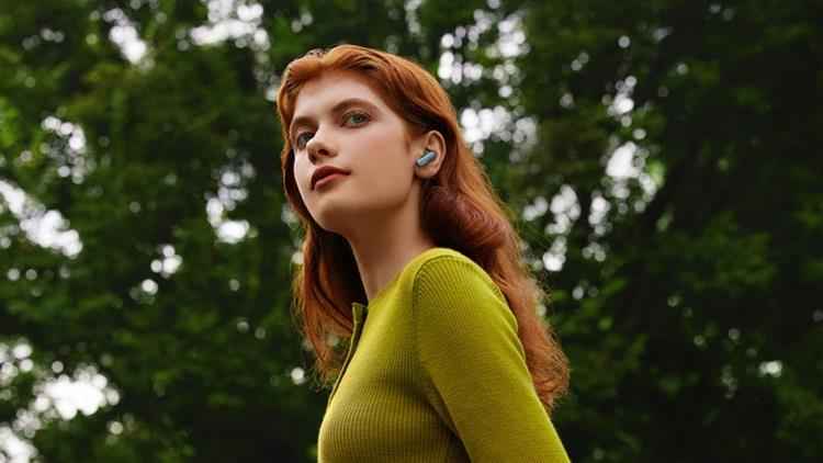 mulher ruiva com fone de ouvido azul, veste blusa verde e está em fundo de plantas desfocadas