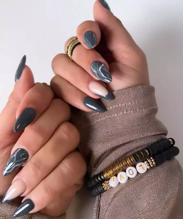 A imagem apresenta mãos com unhas longas e pontiagudas, pintadas em um tom de cinza escuro. Algumas unhas têm um design marmorizado em branco, enquanto outras são lisas. A pessoa usa um suéter bege e pulseiras de miçangas pretas e douradas, com letras formando a palavra "LOVE". O visual é moderno e elegante.


