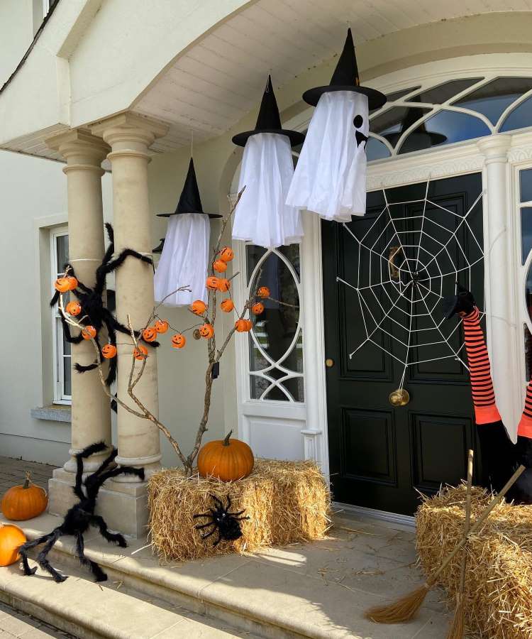 Entrada de casa com parede bege e porta preta com teia de aranha, feno, aranhas gigantes, fantasmas e abóboras