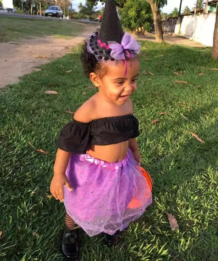 Menina com fantasia de bruxa infantil. Cores predominantes: preto e lilás. Usa: top, saia, meia arrastão, sapatilha, tiara de mini chapéu de bruxa e baldinho em formato de cabeça de abóbora