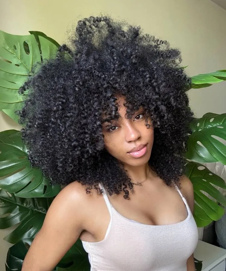 Uma mulher com volumosos cabelos escuros e encaracolados posa contra um fundo de folhas grandes e verdes. Ela usa uma blusa clara e olha suavemente para a câmera.