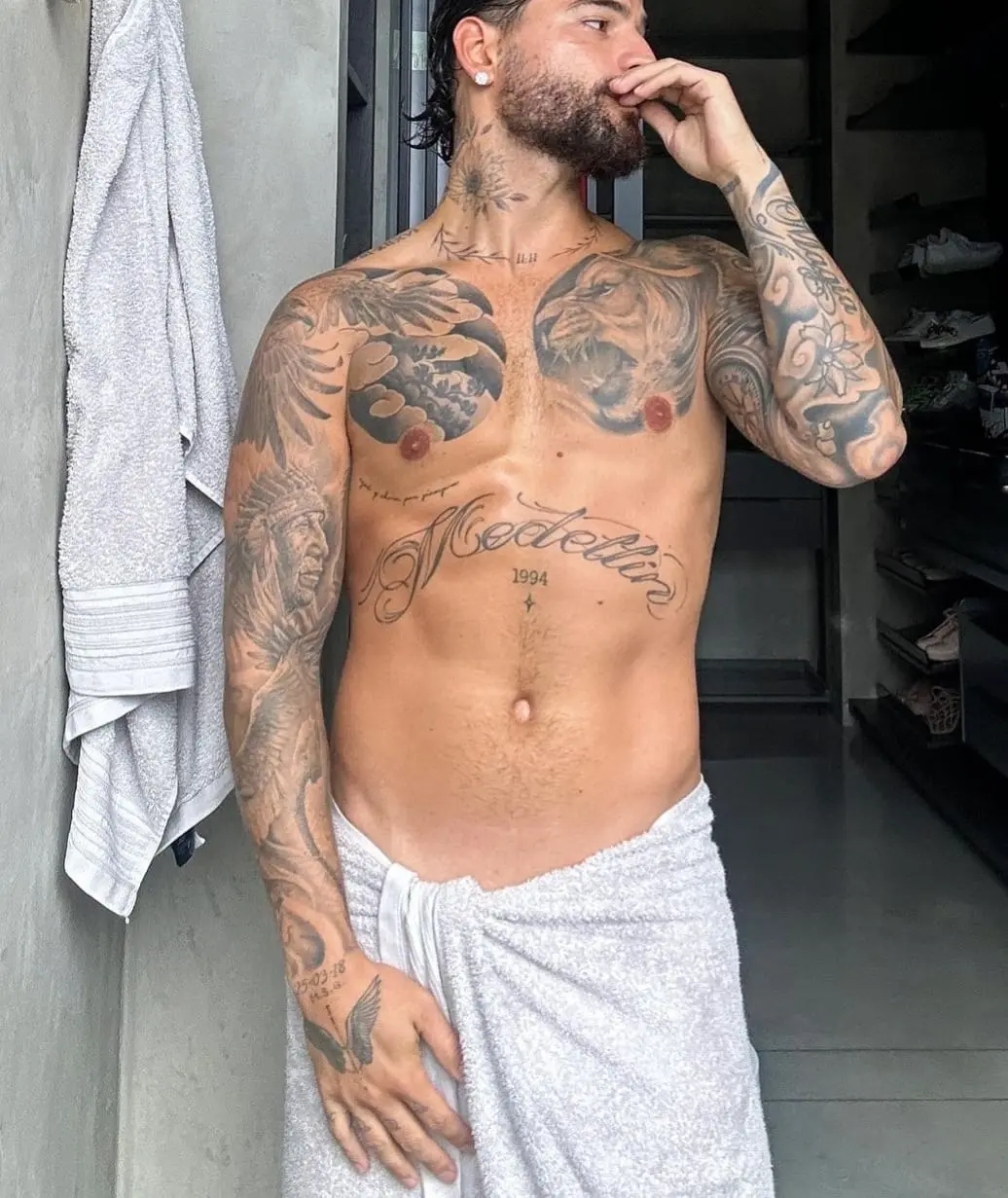 Maluma em varanda com cabelo molhado e toalha amarrada na cintura, exibindo tatuagens no colo, barriga, braços, pescoço e pulsos