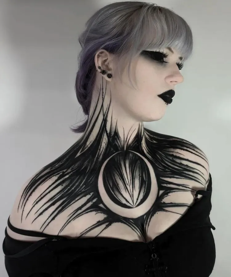 A imagem mostra uma pessoa com cabelo cinza claro e maquiagem escura. Ela tem uma tatuagem no pescoço que se estende até o peito, com um design abstrato em preto, lembrando penas ou garras, e um círculo central formando uma lua. A tatuagem cobre o pescoço e a parte superior do peito.


