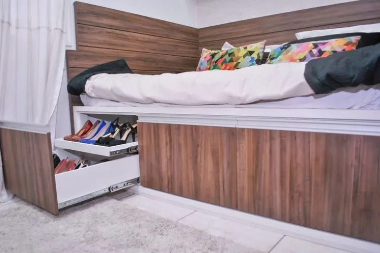 Cama de solteiro na cor marrom com gavetas entre o estrado e o chão, onde foram colocados os calçados. 