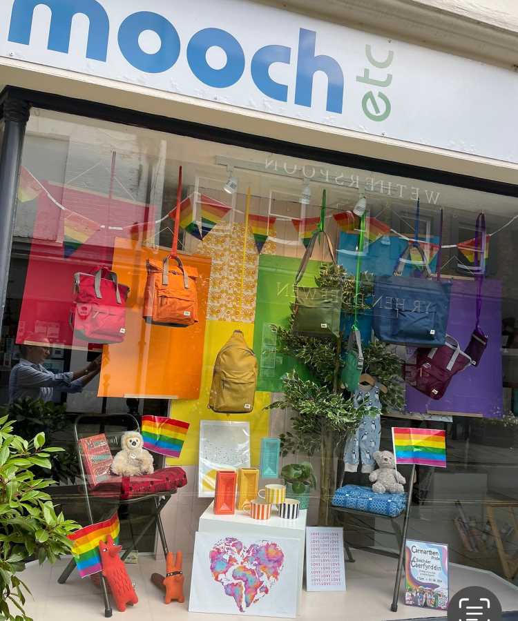 Vitrine colorida com mochilas em tons de arco-íris penduradas. Decoração inclui bandeiras LGBTQIA+, pelúcias, arte em aquarela de coração multicolorido e objetos vibrantes. Plantas completam o espaço vibrante e inclusivo, destacando diversidade e celebração em cenário acolhedor e alegre.