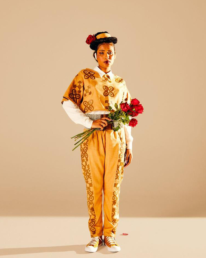 Modelo com blusa e calça amarelas, um chapéu, e segurando flores.