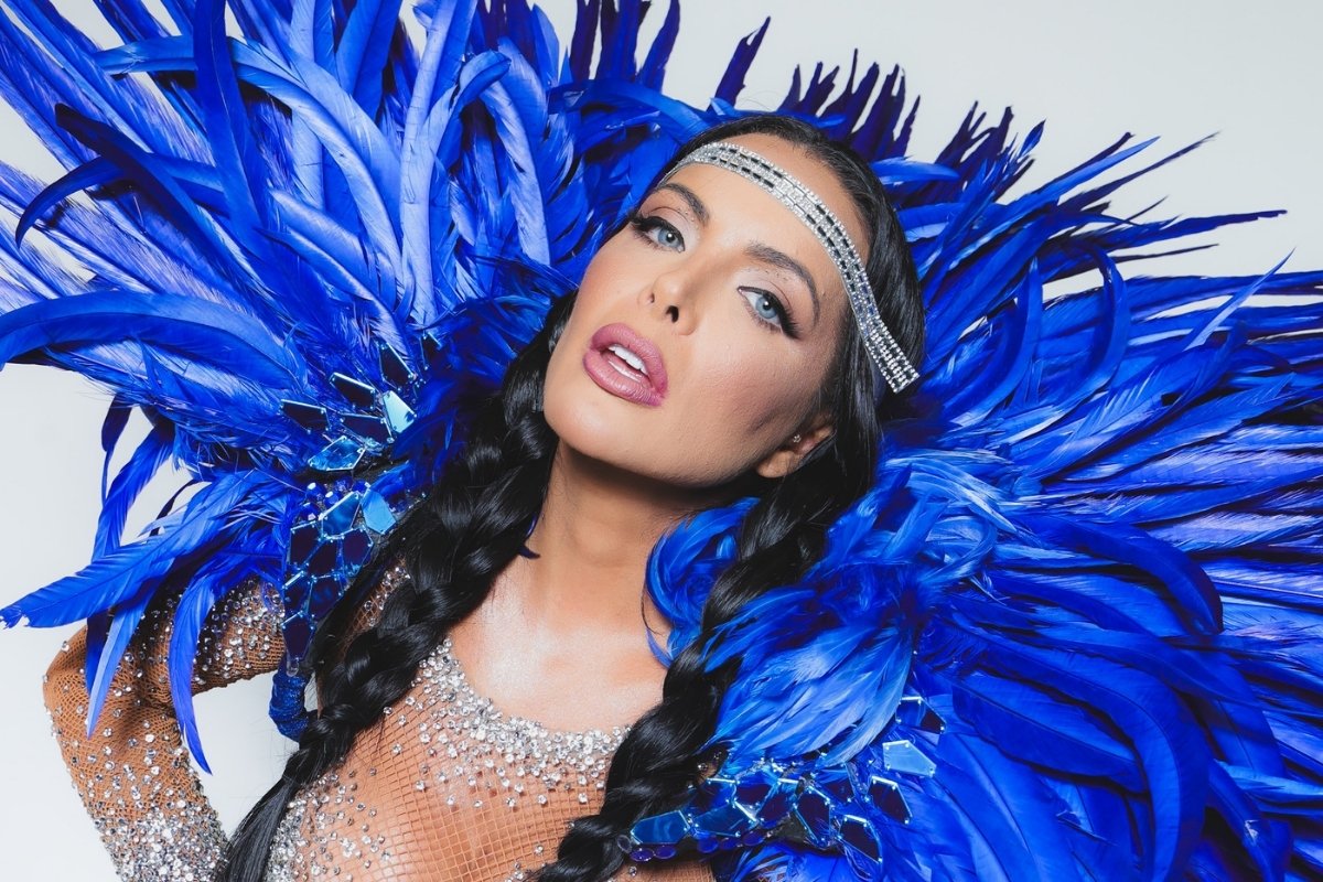 Mulher com cabelo trançado, usando roupa brilhante com pedrarias e elaborada decoração de penas azuis. Detalhe de tiara prateada, maquiagem marcante, olhar intenso, capturada em pose expressiva em um fundo claro.
