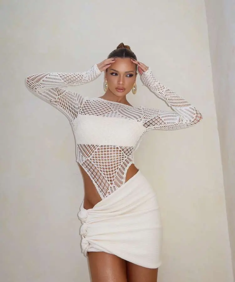 Mulher em vestido branco de crochê, mangas longas, recortes na cintura. Penteado preso, maquiagem suave. Fundo neutro, pose confiante. Acessórios dourados, como brincos, destacam-se. Estilo sofisticado e moderno.