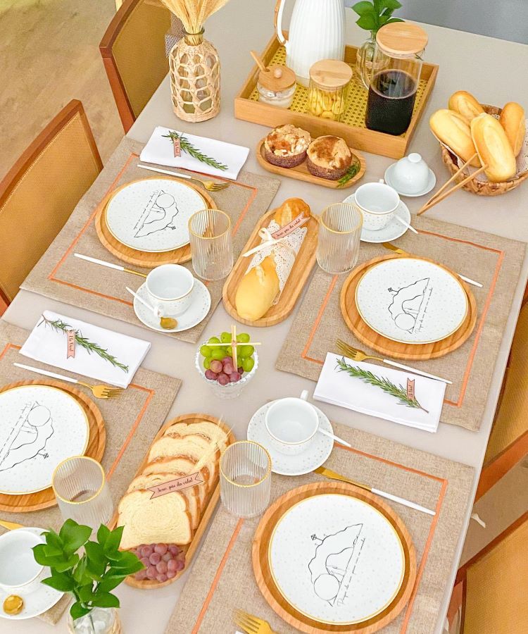 Mesa posta com toalhas bege, pratos com ilustrações religiosas, guardanapos decorativos com raminhos verdes, taças de vidro, xícaras brancas com detalhes dourados, pães artesanais, uvas, mini muffins, jarra de café, garrafa decorativa e arranjo com flores secas.