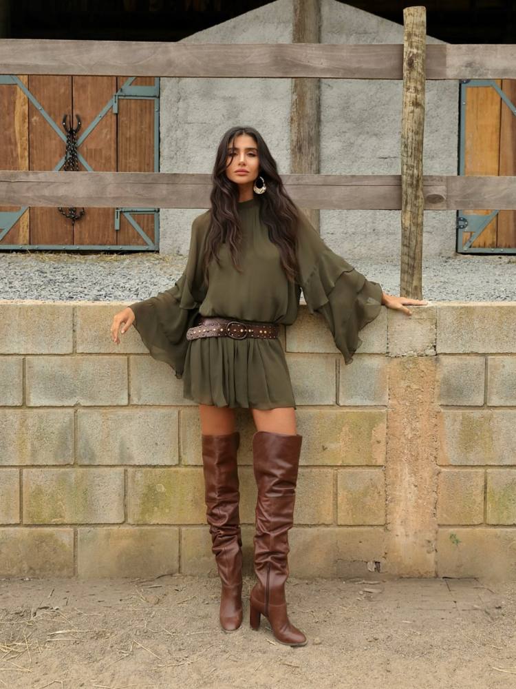 Mulher de pele clara usando vestido curto verde fluido com cinto e bota marrom cano alto 