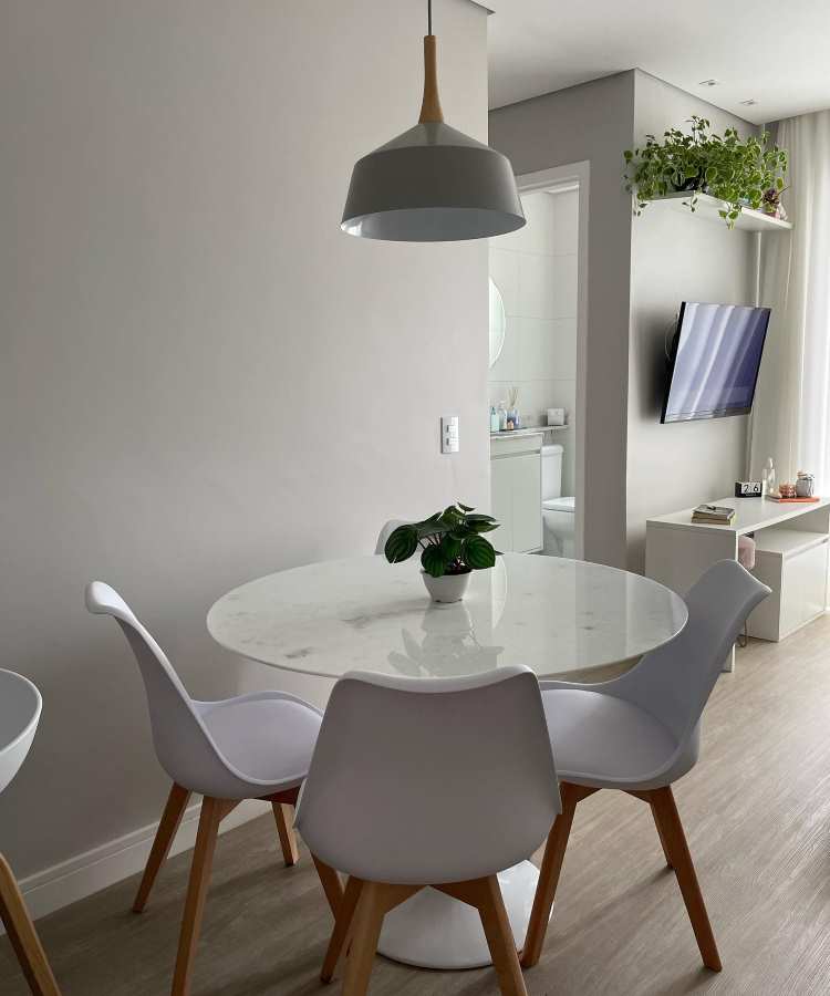 Sala de jantar moderna com mesa redonda de mármore branco, cercada por cadeiras brancas com pés de madeira. No centro da mesa, há uma Peperômia Melancia em vaso branco. Ao fundo, estante, TV e planta suspensa.