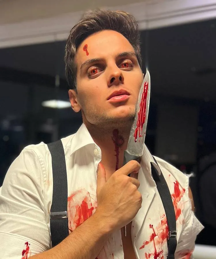 Homem de pele clara usando lente vermelha, sangue falso pelo rosto e pescoço, faca com sangue falso e roupa ensaguentada 