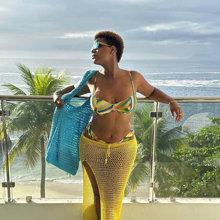 Mulher com cabelo curto e óculos de sol, posando em uma varanda com vista para o mar. Ela usa um biquíni listrado colorido e uma longa saída de praia de crochê amarela com fenda.