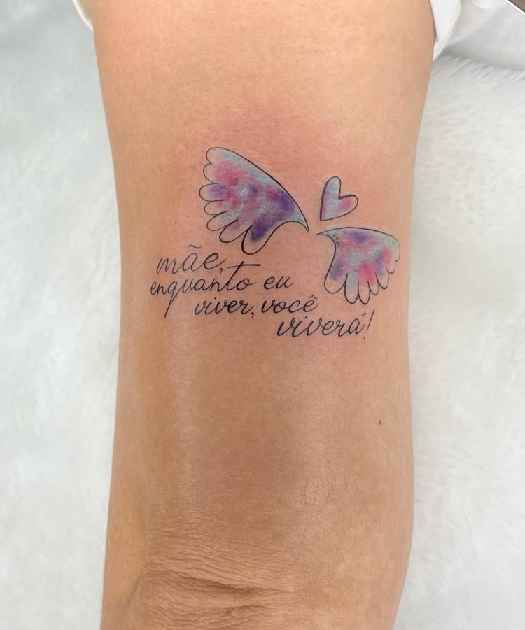 Uma tatuagem delicada no antebraço de uma pessoa. Inclui um par de asas de anjo com tons suaves de roxo e rosa, um pequeno coração e a frase "mãe, enquanto eu viver, você viverá!" em caligrafia.