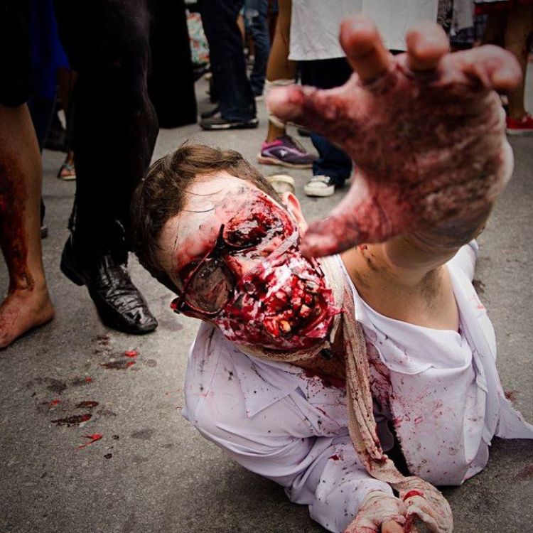 Homem de pele clara no chão usando roupa branca e maquiagem zumbi com muito sangue falso 