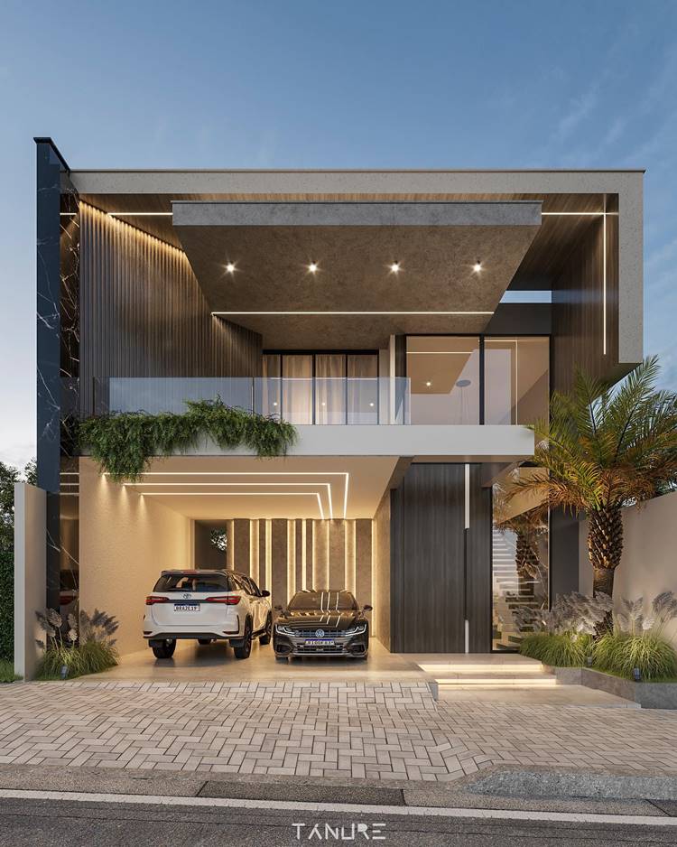A imagem mostra uma casa moderna, com dois carros na garagem, iluminação embutida e jardim na frente. A casa tem uma combinação de texturas de concreto e madeira. O andar superior tem uma varanda com uma grade de vidro e é parcialmente coberto. Há várias plantas e pequenas árvores ao redor da entrada da casa, adicionando verde à arquitetura moderna.