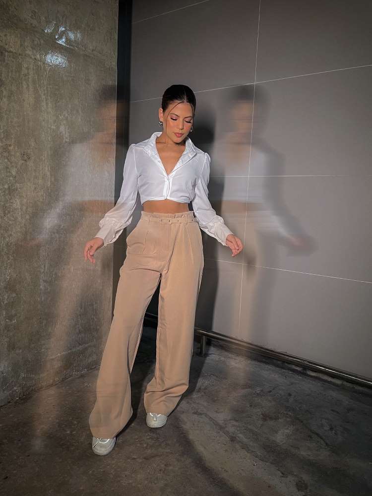 Mulher de pele clara usando camisa branca como cropped e calça marrom de alfaiataria. 
