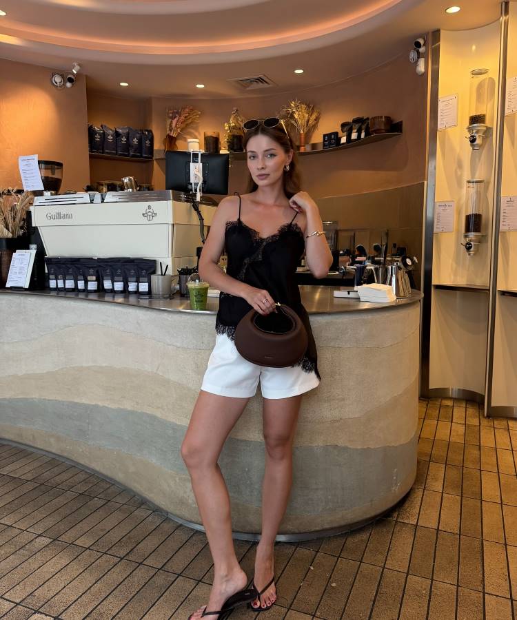 Uma mulher posa casualmente em uma cafeteria. Ela veste uma blusa de alças preta com renda, shorts brancos e chinelos. Ela segura uma bolsa marrom e usa óculos de sol na cabeça, encostada no balcão da loja.