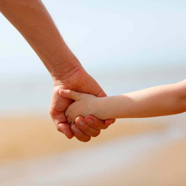 Duas mãos se seguram com carinho, um adulto e uma criança, simbolizando proteção, cuidado e conexão. Ao fundo, desfocado, aparece uma praia com tons suaves de areia e céu azul, transmitindo tranquilidade e amor.