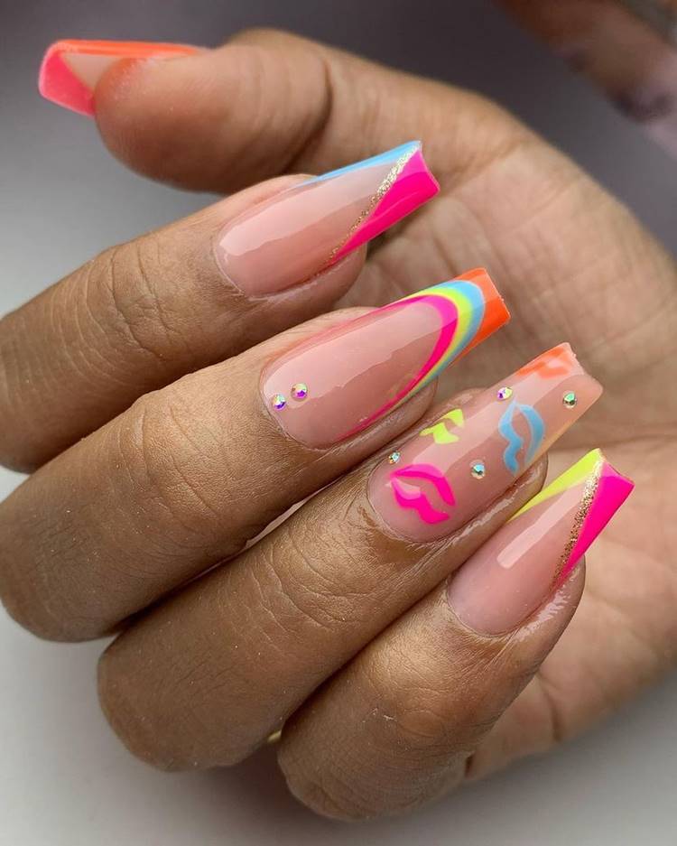 Mão com unhas arco-íris LGBTQIA+: Polegar: decoração em V pink e laranja neon com risco dourado Indicador: decoração em V pink e azul neon com risco dourado Médio: arco-íris na franceinha sorrindo com 2 pedrinhas na cutícula Anelar: boquinhas coloridas com pedrinhas brilhantes Mínimo decoração em V pink e amarelo neon com risco dourado 