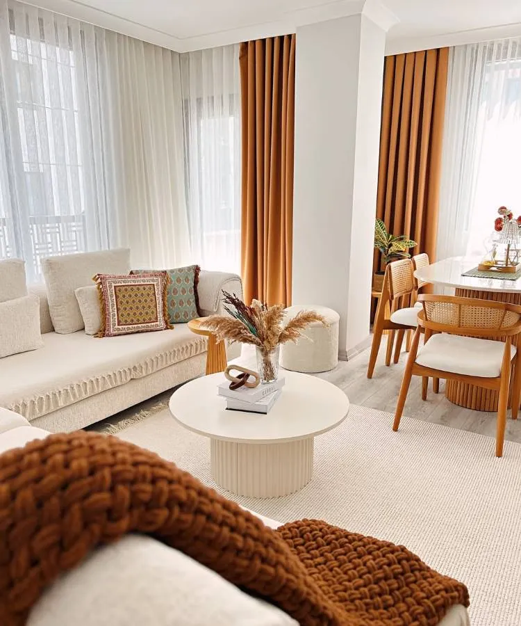 Uma sala de estar e jantar integrada, com sofá creme e almofadas para sala estampadas. A mesa de centro redonda exibe um arranjo floral. Cortinas em tons terrosos complementam a área de jantar com mesa branca e cadeiras de madeira. O ambiente é convidativo e harmonioso.








