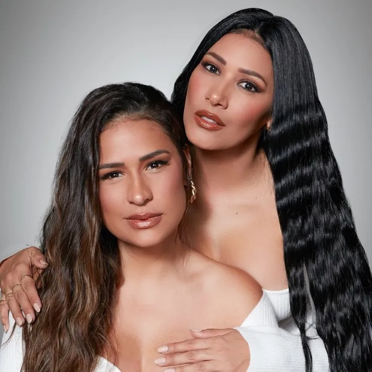 Simone & Simaria  posam juntas contra um fundo cinza suave. A da esquerda tem pele média e cabelo castanho ondulado. A da direita, pele mais clara e cabelo preto liso. Ambas usam roupas brancas elegantes. O ambiente é íntimo e sereno.