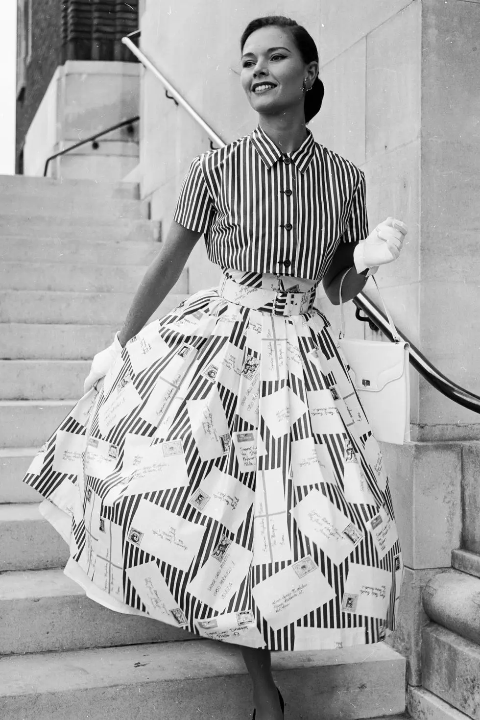 Modelo a posar com uma saia e camisa listradas nos anos 50, com luvas brancas nas mãos. 