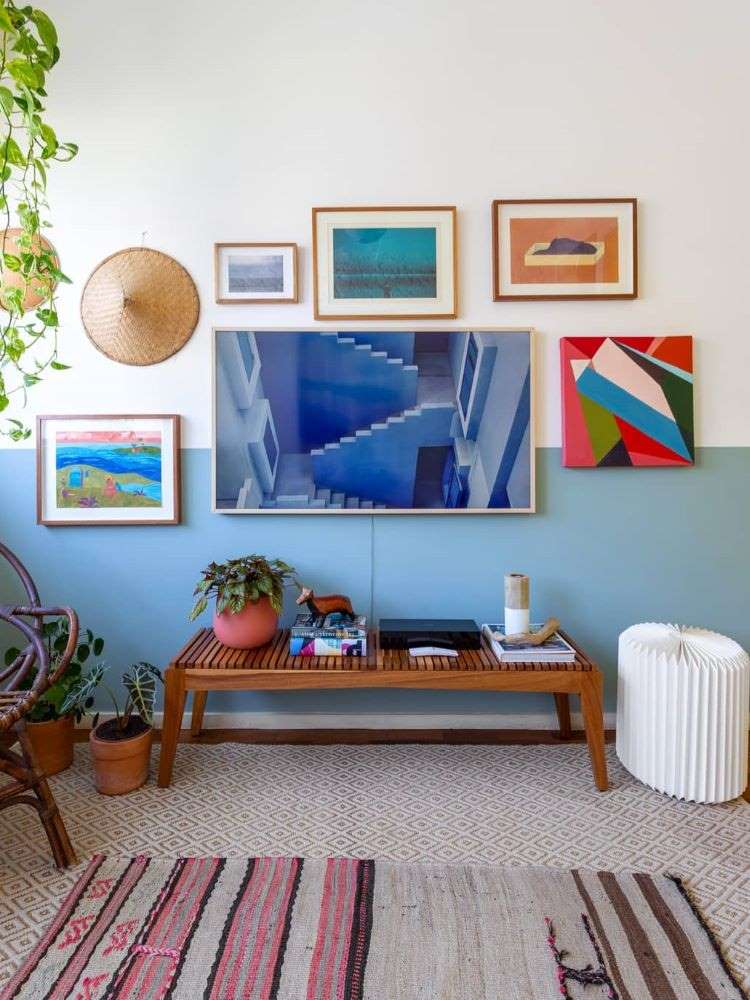 Sala de estar com móvel em madeira, vasos, meia parede azul e vários quadros pregados, dentre eles uma televisão que imita quadro quando desligada.