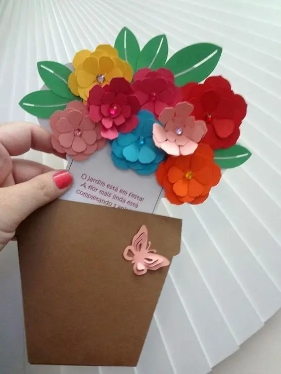 Pessoa de pele clara usando esmalte vermelho segurando cartão com flores coloridas e vaso com borboleta de papel