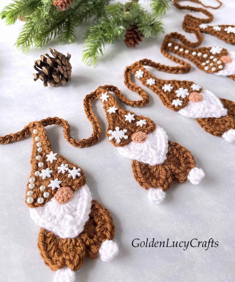 Adoráveis gnomos de crochê, decorados com pérolas e flocos de neve, estão alinhados sobre uma superfície clara. Ao fundo, um ramo de pinheiro e uma pinha complementam o tema natalino artesanal.


