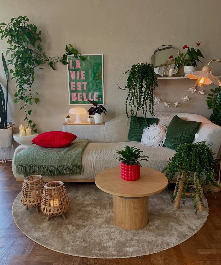Uma sala de estar com sofá bege, almofadas verdes e uma manta verde. Plantas em vasos e penduradas decoram o ambiente, com uma mesa de centro redonda de madeira, luminárias de vime e um quadro "La Vie Est Belle" na parede.