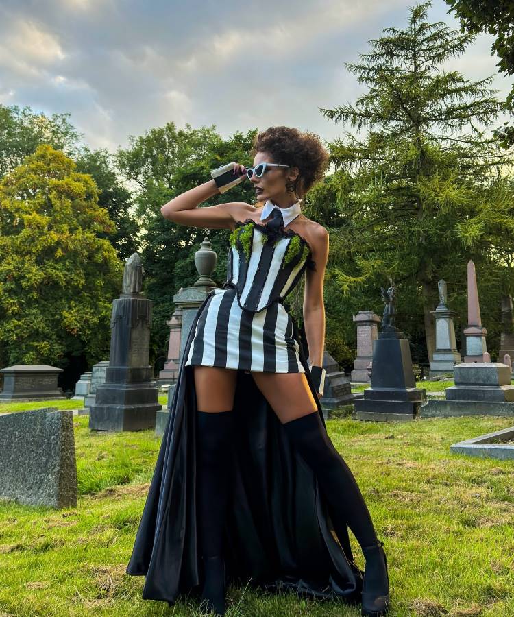 A imagem mostra uma pessoa em um cemitério, vestindo uma fantasia com listras verticais em preto e branco. O traje inclui um corpete e uma saia curta, complementados por uma longa capa preta. Ela usa óculos escuros e luvas pretas, posando de forma confiante com uma mão na cintura. O cenário ao fundo é composto por lápides e árvores, criando um ambiente dramático e misterioso. A iluminação suave do céu nublado adiciona um toque melancólico à cena.


