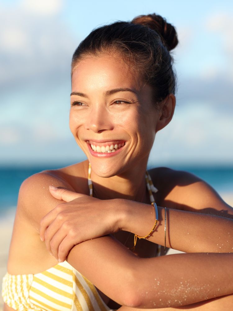 Uma mulher sorridente com cabelos castanhos presos em um coque. Ela está usando uma blusa de alças listrada amarela e branca, sentada na areia da praia, com o mar azul ao fundo.