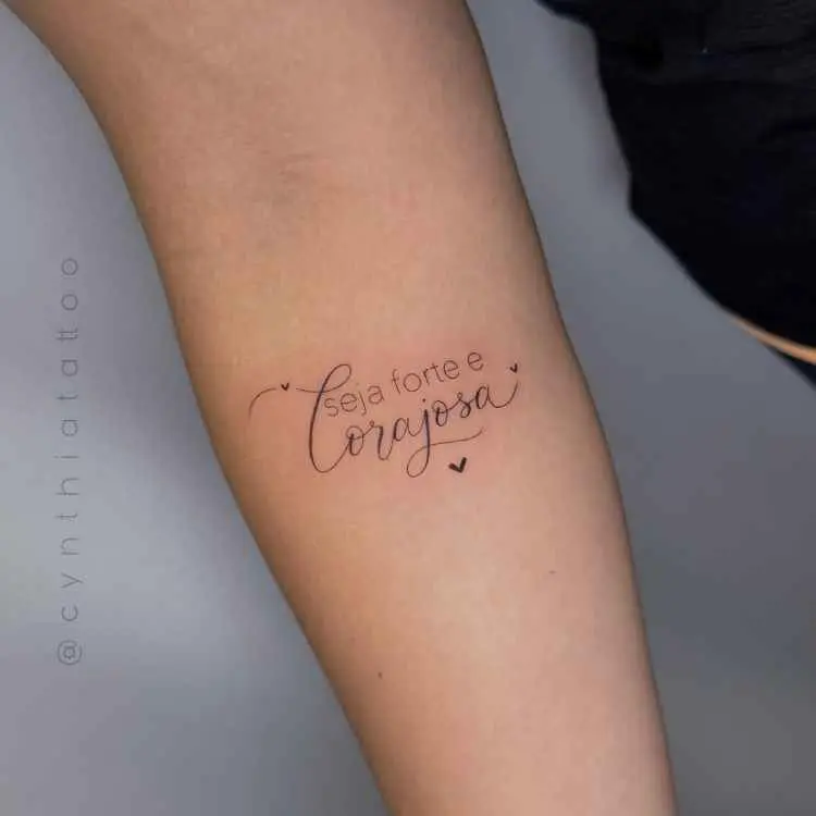 A imagem mostra uma tatuagem no braço com a frase delicada: "seja forte e corajosa", combinando fontes simples e cursivas. Há pequenos corações ao redor, dando um toque feminino e motivador. O fundo é neutro, destacando a tatuagem.