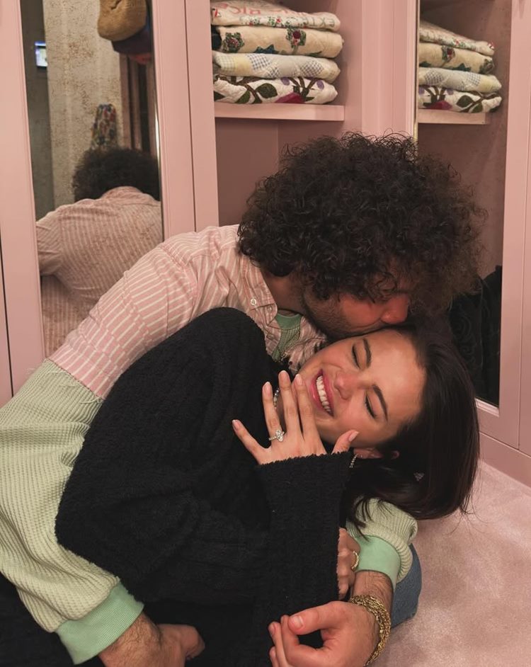 Um homem de cabelos cacheados e barba beija a testa de uma mulher sorridente que está aninhada em seu colo. A mulher exibe um anel brilhante na mão esquerda, parecendo feliz e relaxada.