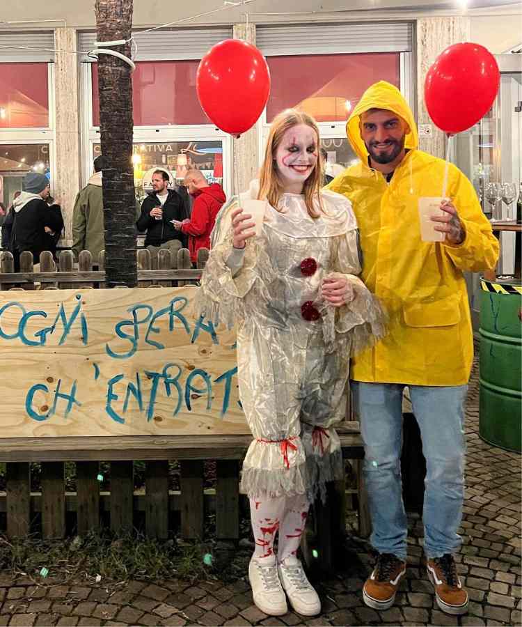 Fantasia de casal para o Halloween: Pennywise e Georgie Denbrough de "It: A Coisa". A mulher está vestida com roupa de palhaço branca e ele usa calça jeans e capa de chuva amarela. Ambos segurando bexiga vermelha 