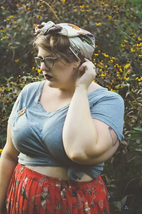 Frase para foto pensativa plus size usa lenço na cabeça, óculos de grau, batom vinho e veste saia plissada estampada com t-shirt cinza Frase para foto pensativa plus size