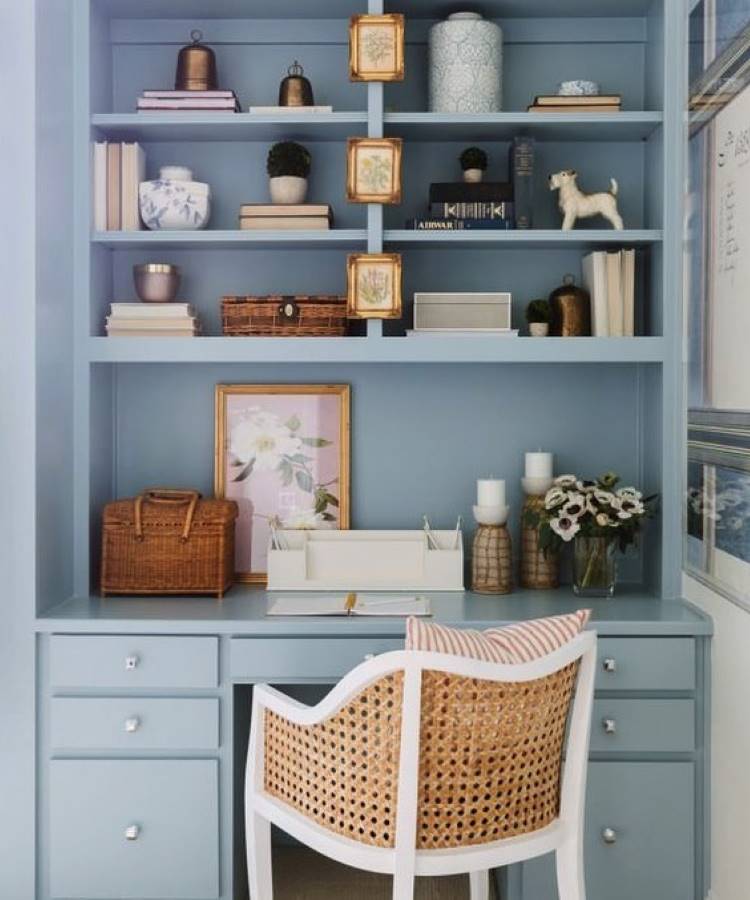 O ambiente apresenta uma escrivaninha azul claro com gavetas e prateleiras decoradas com livros, vasos, cestos e quadros. Uma cadeira branca com encosto de palha acompanha o espaço. Detalhes como flores, velas e molduras douradas trazem charme e elegância.