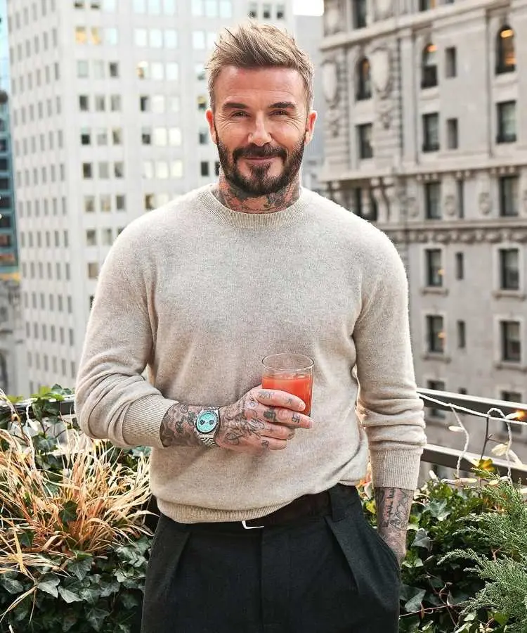 A imagem mostra David Beckham, um dos influencers de moda masculina, sorridente, vestindo um suéter bege e calça preta, segurando um copo de bebida vermelha. Ele exibe tatuagens nos braços e usa um relógio prateado, posicionado em uma varanda com prédios ao fundo.
