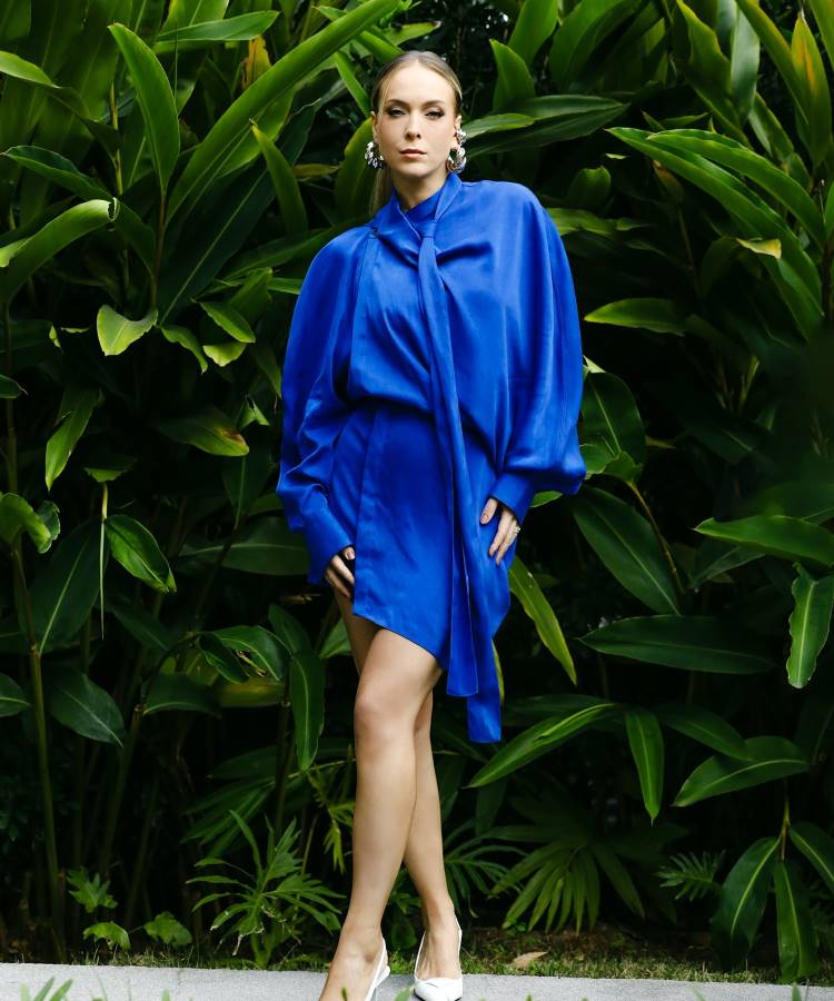 A imagem apresenta uma mulher posando em frente a uma folhagem verde exuberante. Ela veste um vestido azul-royal de seda com mangas bufantes e gola alta com laço. Nos pés, sapatos brancos de salto e, nas orelhas, brincos grandes e prateados.