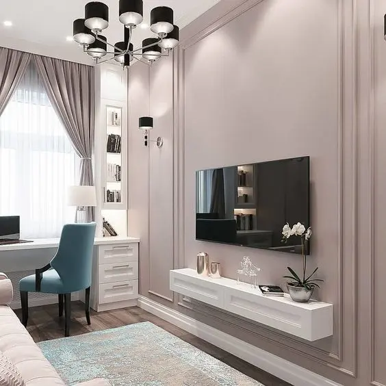 Home office com TV em parede rosa antigo com moldura, aparador branco suspenso com itens decorativos, mesa branca, cadeira azul, estante de canto branca iluminada