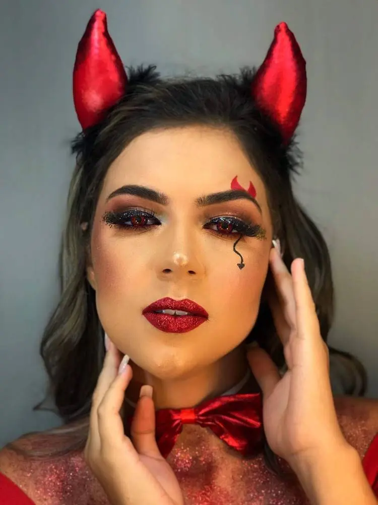 Mulher de pele clara usando tiara vermelha de diabo, delineado preto, desenho de chifre sobre a sobrancelha e rabo de diabo abaixo dos olhos, além de batom vermelho brilhante