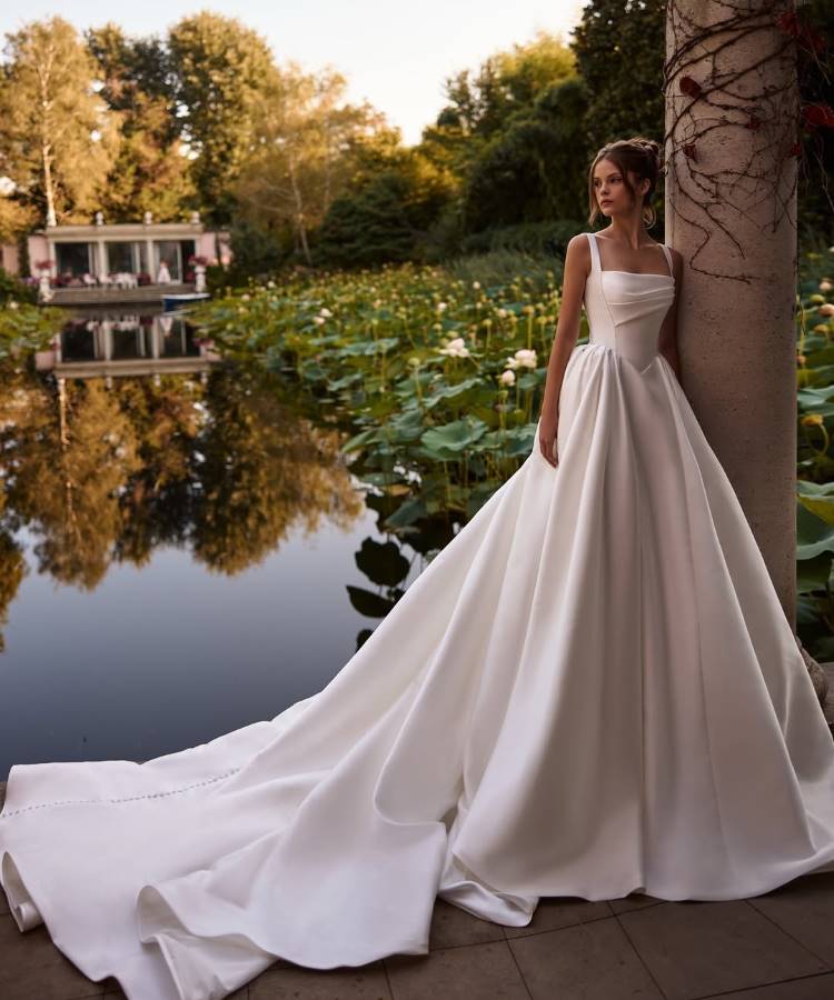 Noiva em vestido branco minimalista com alças finas, decote quadrado e saia volumosa com longa cauda, refletindo as tendências em vestidos de noiva 2025. O cenário natural com lago, flores de lótus e vegetação cria um ambiente romântico.





