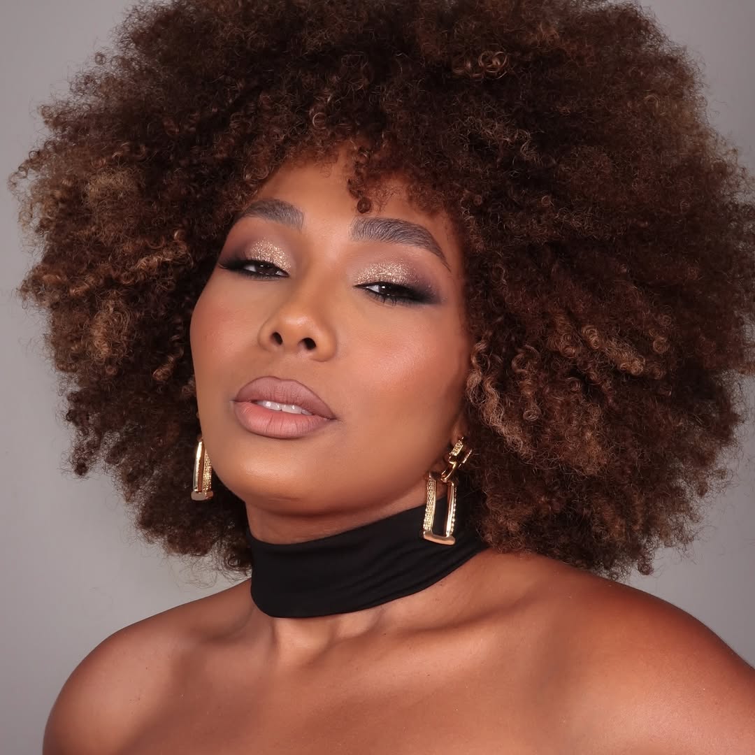 Mulher com cabelos crespos volumosos em tons de castanho iluminado. Ela usa maquiagem sofisticada com sombras brilhantes, batom nude e tons terrosos. Veste acessórios dourados e uma faixa preta no pescoço, destacando sua expressão confiante e elegante.