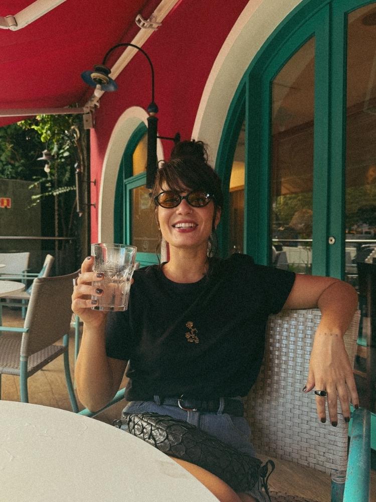 Thais de blusa preta sentada à mesa tomando um copo de água 