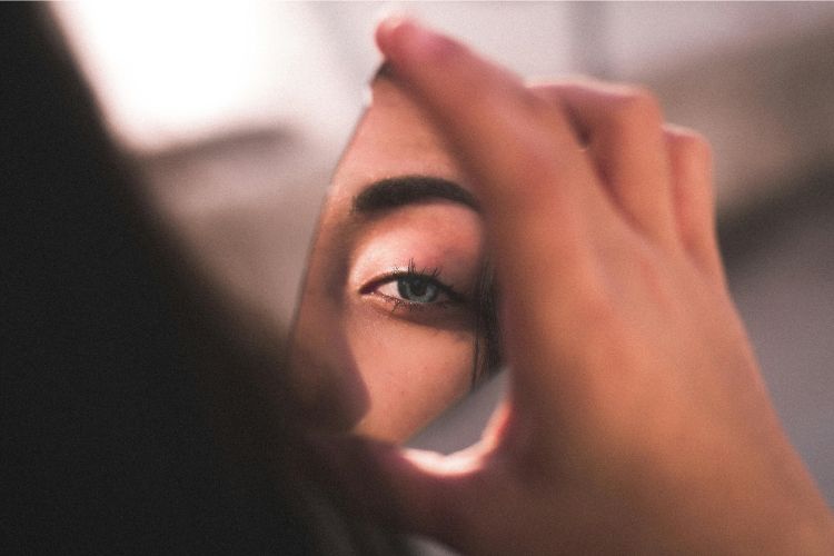 Foto de mulher de olho azul e cabelo castanho segurando um caco de espelho e observando seu reflexo